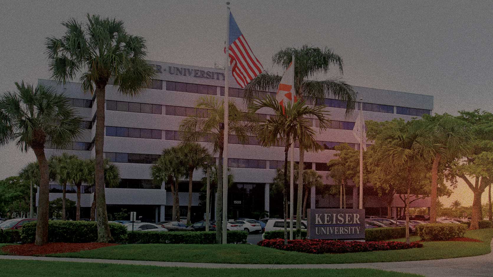 Keiser University campus building with palm trees, flags, and parked cars under a partly cloudy sky.