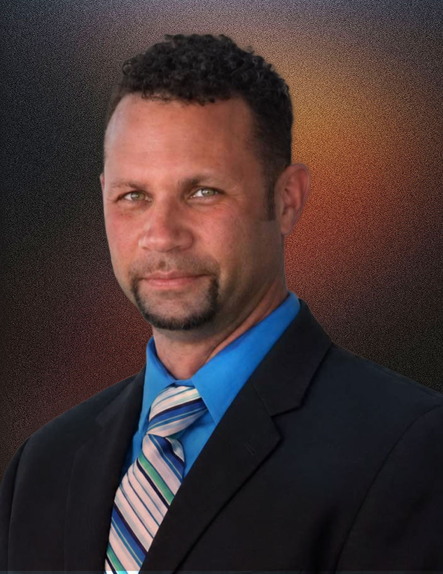 Man with short curly hair and goatee wearing a black suit jacket, blue dress shirt, and striped blue tie, standing indoors in front of windows.
