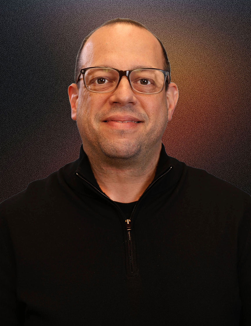 Man with short curly dark hair and glasses, wearing a navy blue quarter-zip pullover, smiling slightly in a modern office with large windows and plants in the background.