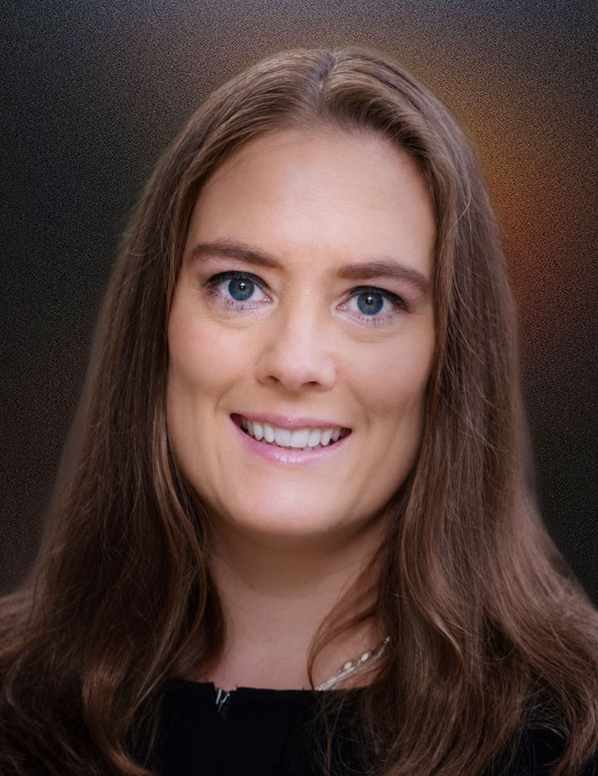 Portrait of a woman with long brown hair and blue eyes smiling against a blurred outdoor background.