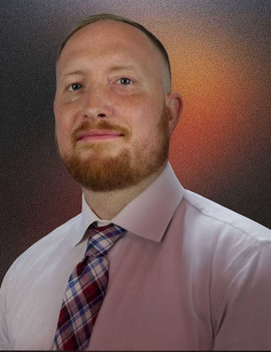 Portrait of a man with short light brown hair and a beard wearing a white shirt and a red, blue, and white plaid tie, against a gray background.