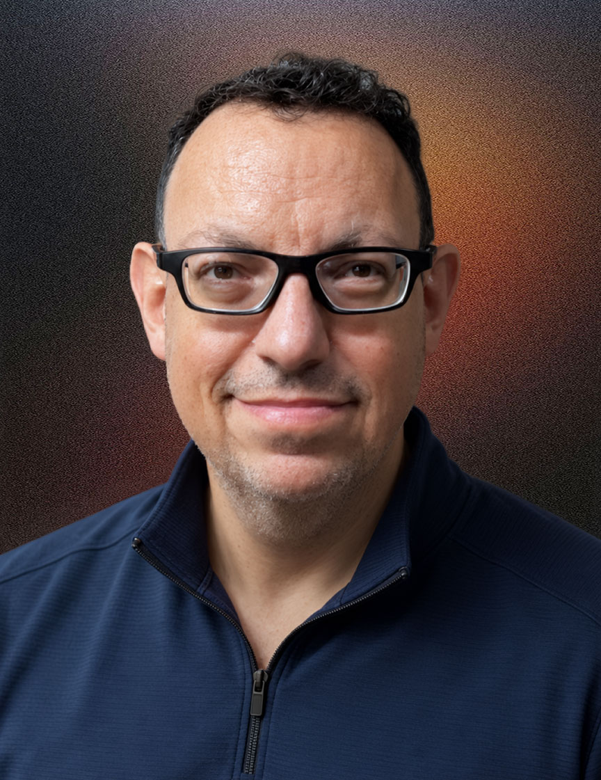 Man with short curly dark hair and glasses, wearing a navy blue quarter-zip pullover, smiling slightly in a modern office with large windows and plants in the background.