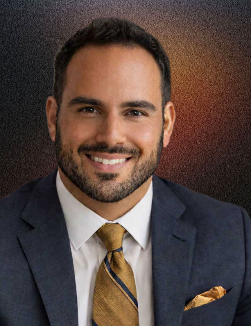 Smiling man with short dark hair and beard wearing a dark blue suit, white shirt, and golden striped tie, seated in an office with books and a lamp in the background.