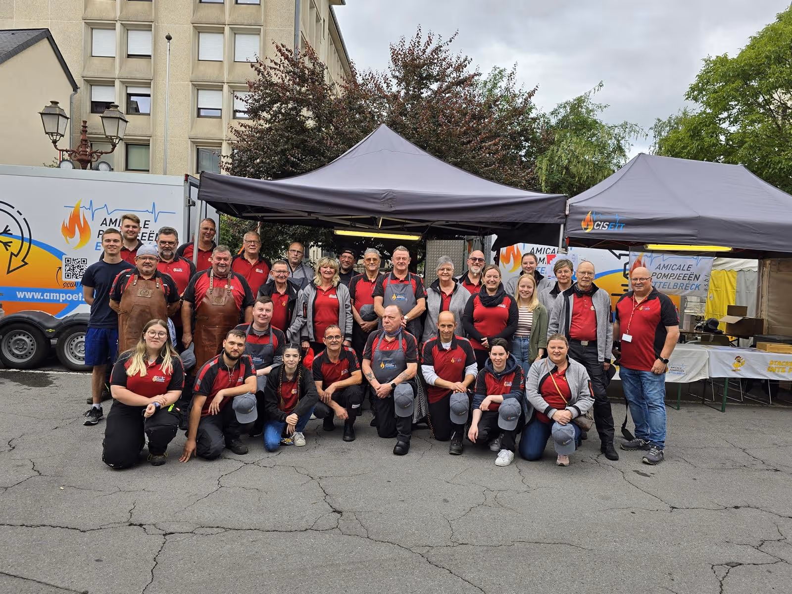 Group of people in red shirts posing outdoors under gray tents with event trailers in background.