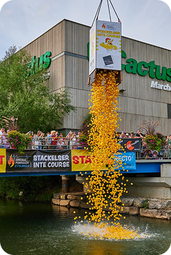 Large box pouring thousands of yellow balls from a bridge into water below with spectators watching.