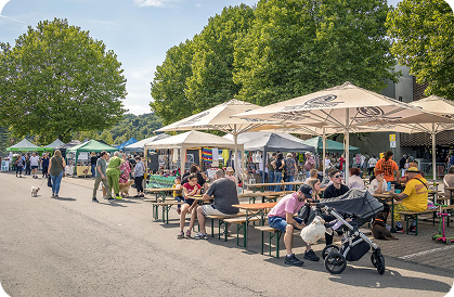 Outdoor market with people sitting under large umbrellas and walking among vendor tents on a sunny day.