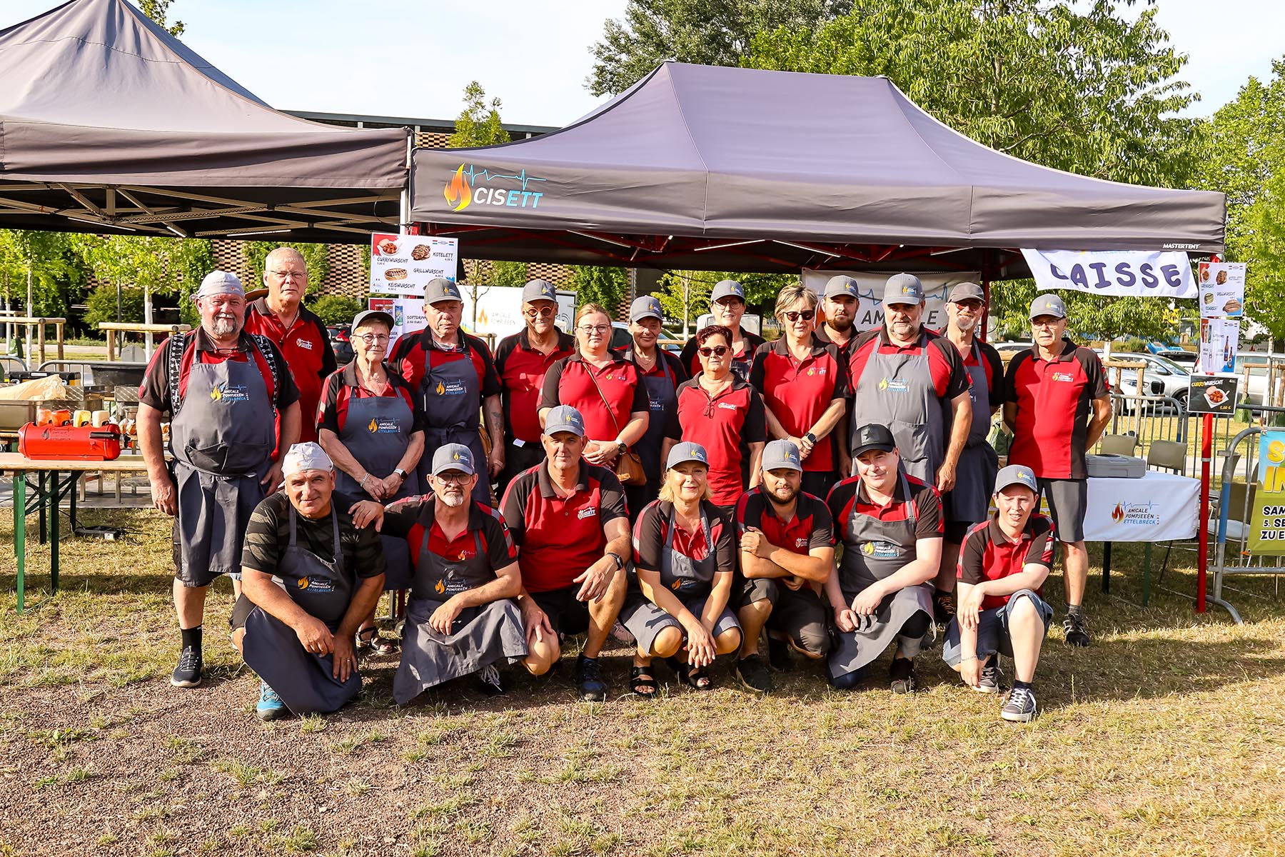 Group of people in red shirts posing outdoors under gray tents with event trailers in background.