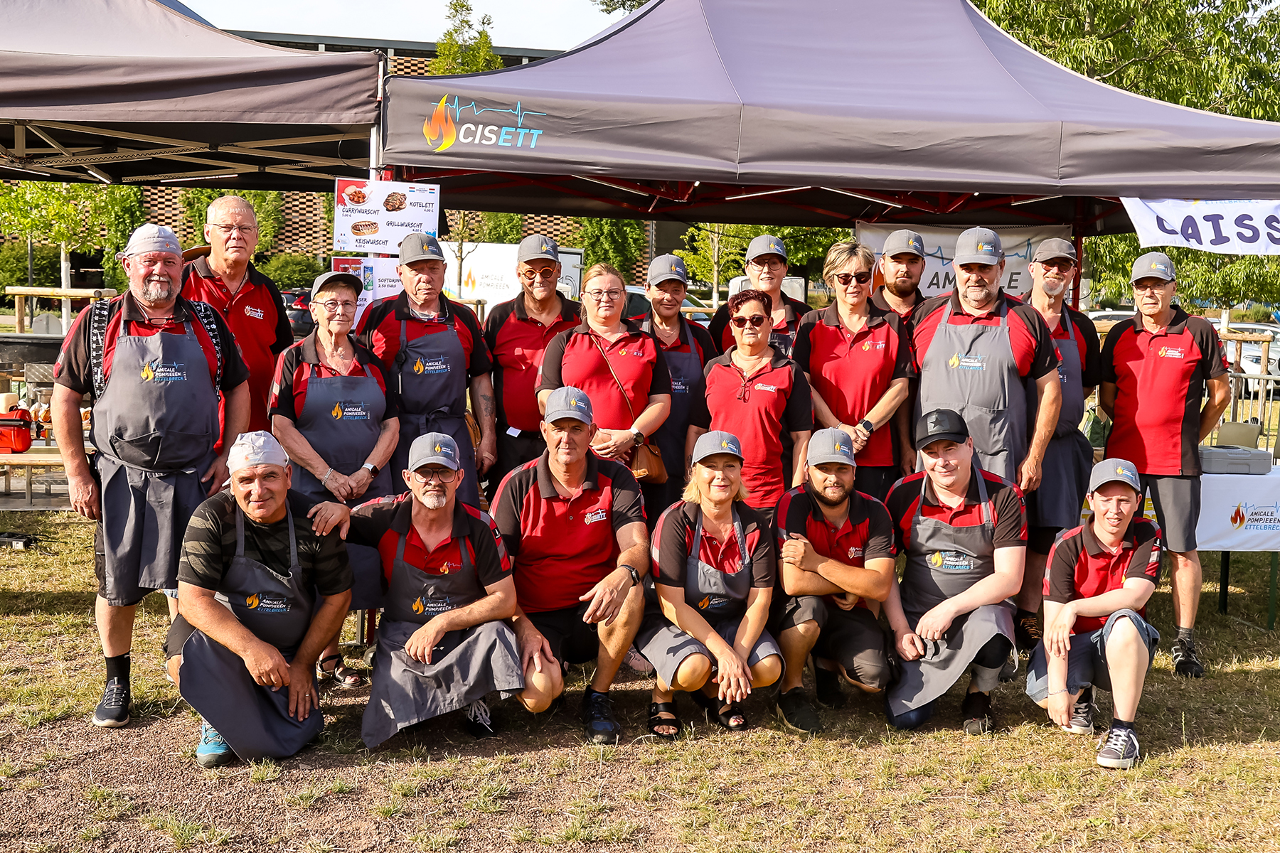 Group of people in red shirts posing outdoors under gray tents with event trailers in background.