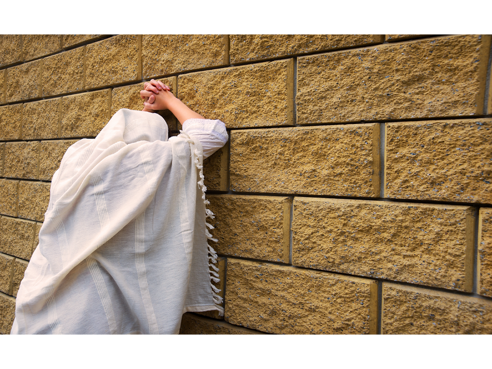 Person in white shawl praying at a textured stone wall, arms raised and crossed. The setting is calm, with earthy tones and no visible text.