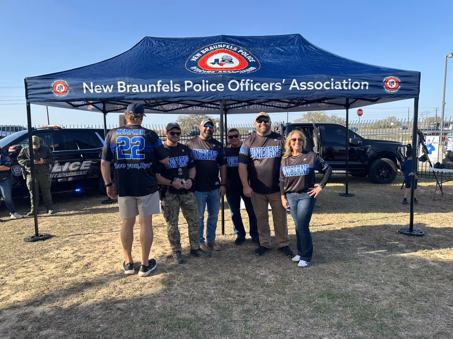 Group of six people under a New Braunfels Police Officers' Association tent wearing matching black Unicorns shirts.