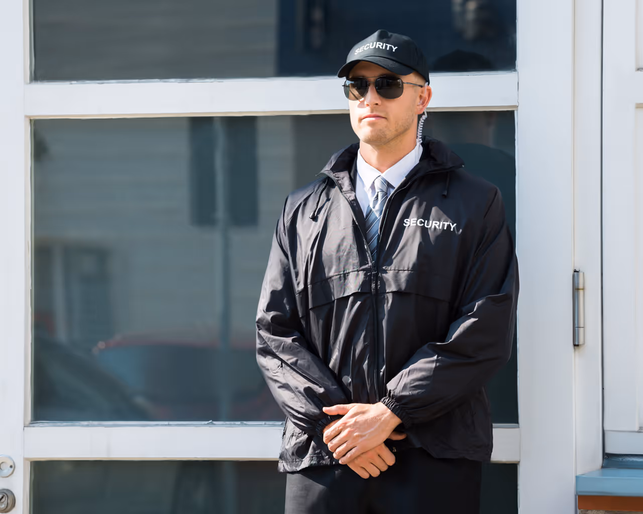 Security guard in sunglasses and black jacket standing with hands clasped in front of a building entrance.