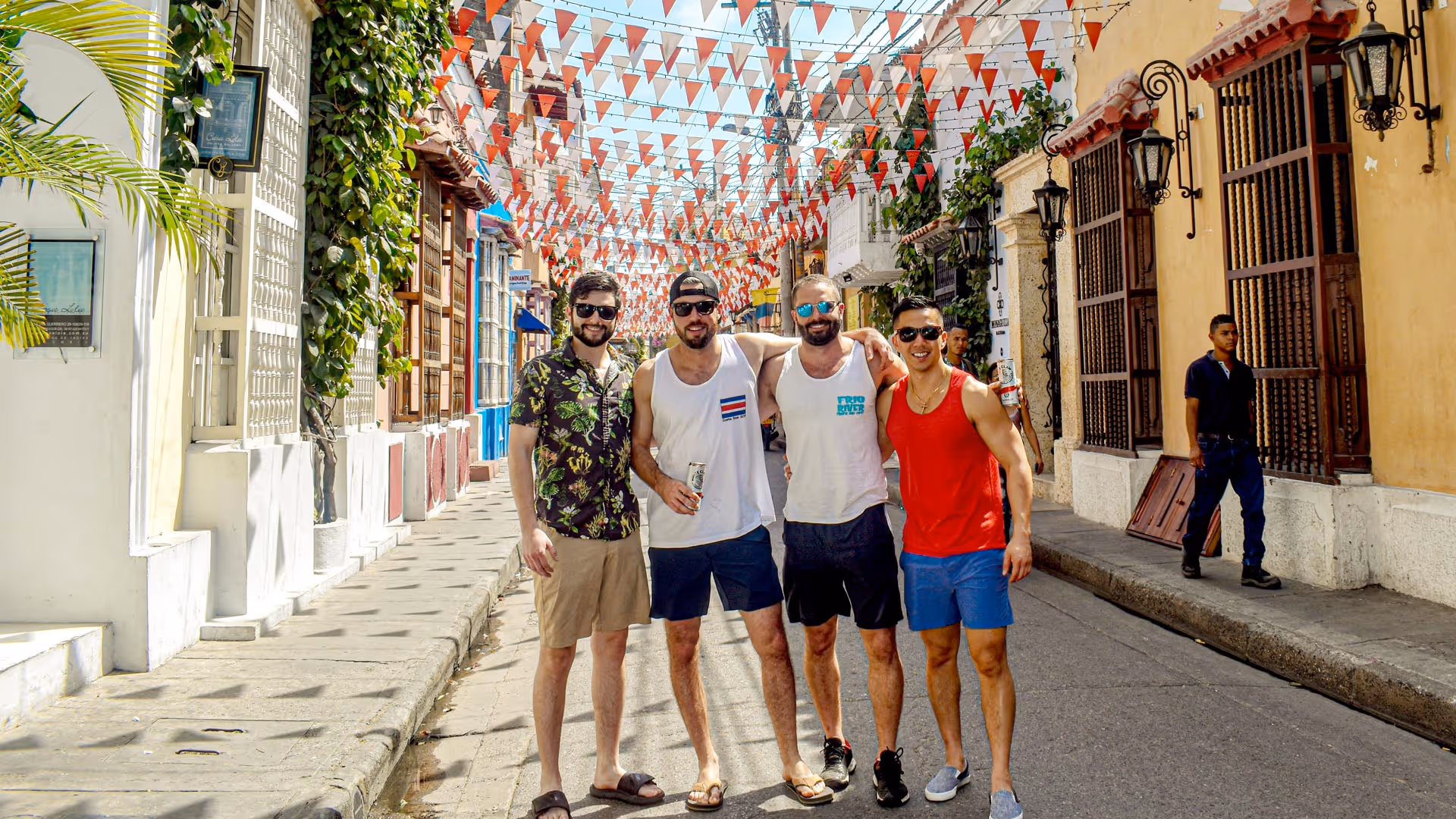 Four men wearing summer clothes and sunglasses stand close on a festive street decorated with red and white flags.