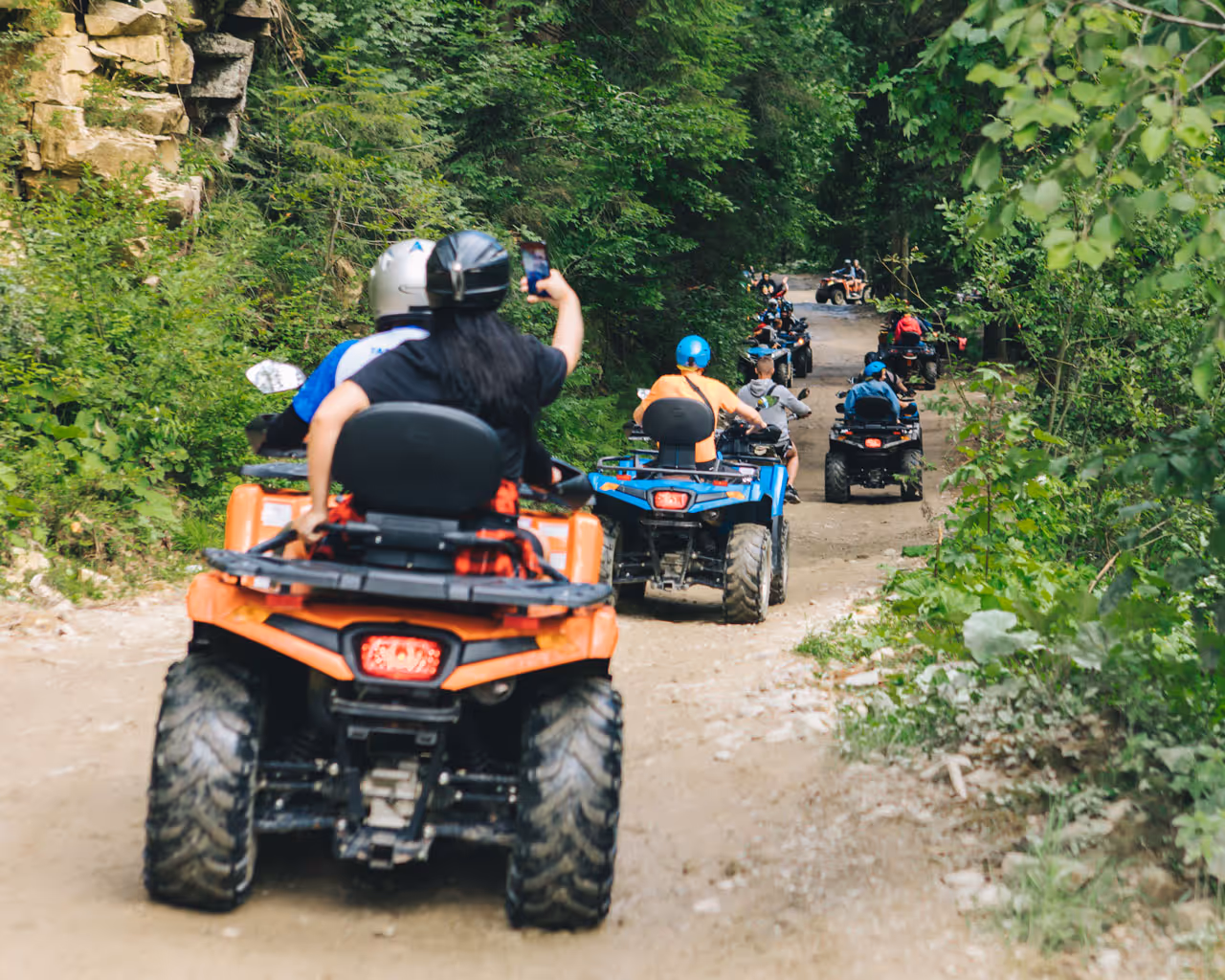 Group riding ATVs on a forest dirt trail surrounded by green trees and rocky terrain.