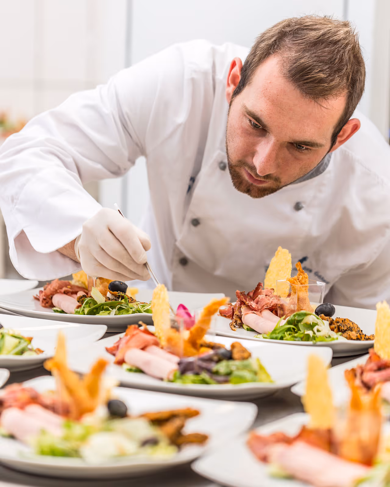 Chef in white uniform carefully plating gourmet dishes with meats and greens on white plates.