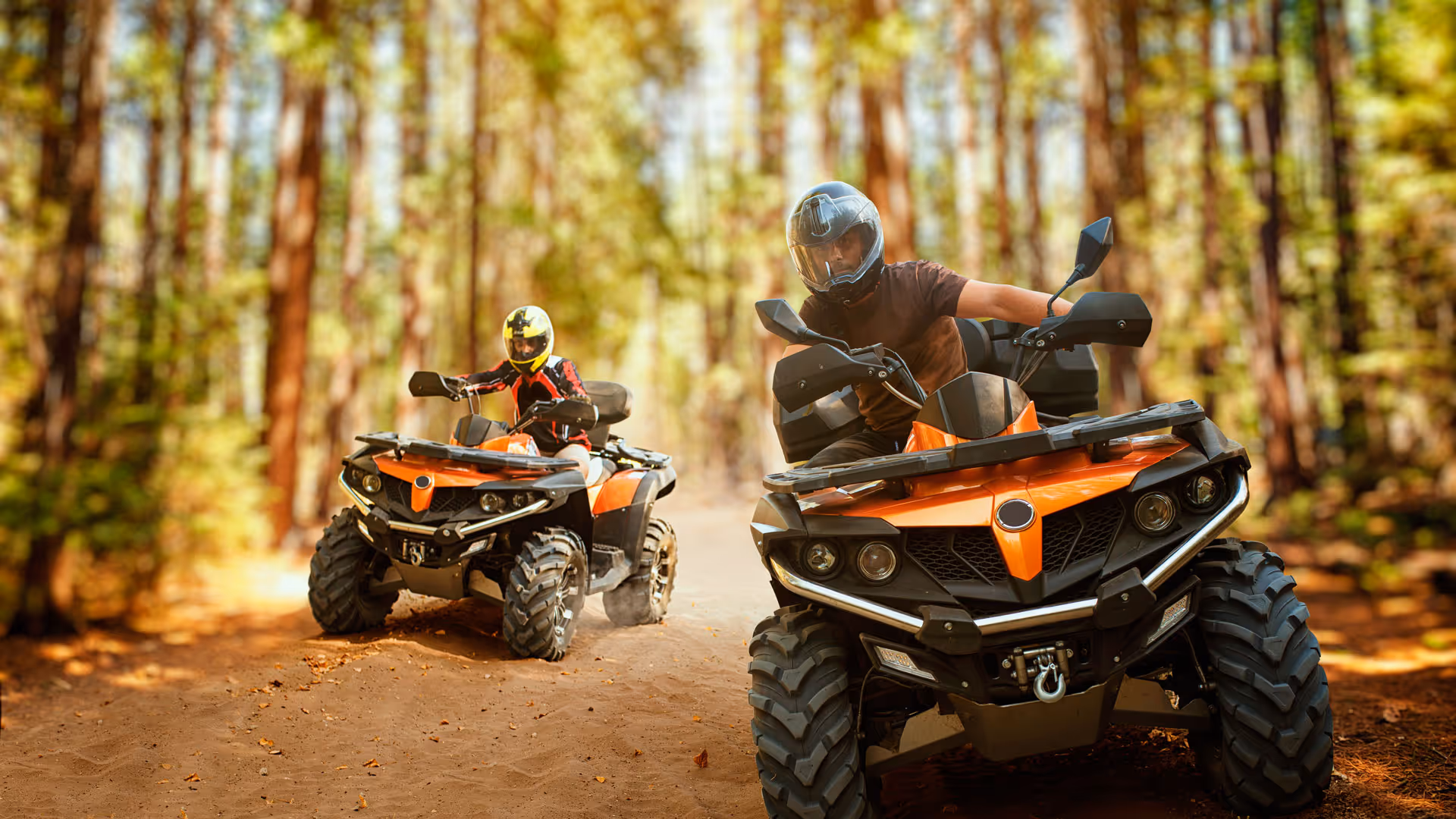 Two riders wearing helmets driving orange ATVs on a forest trail covered with dirt and leaves.