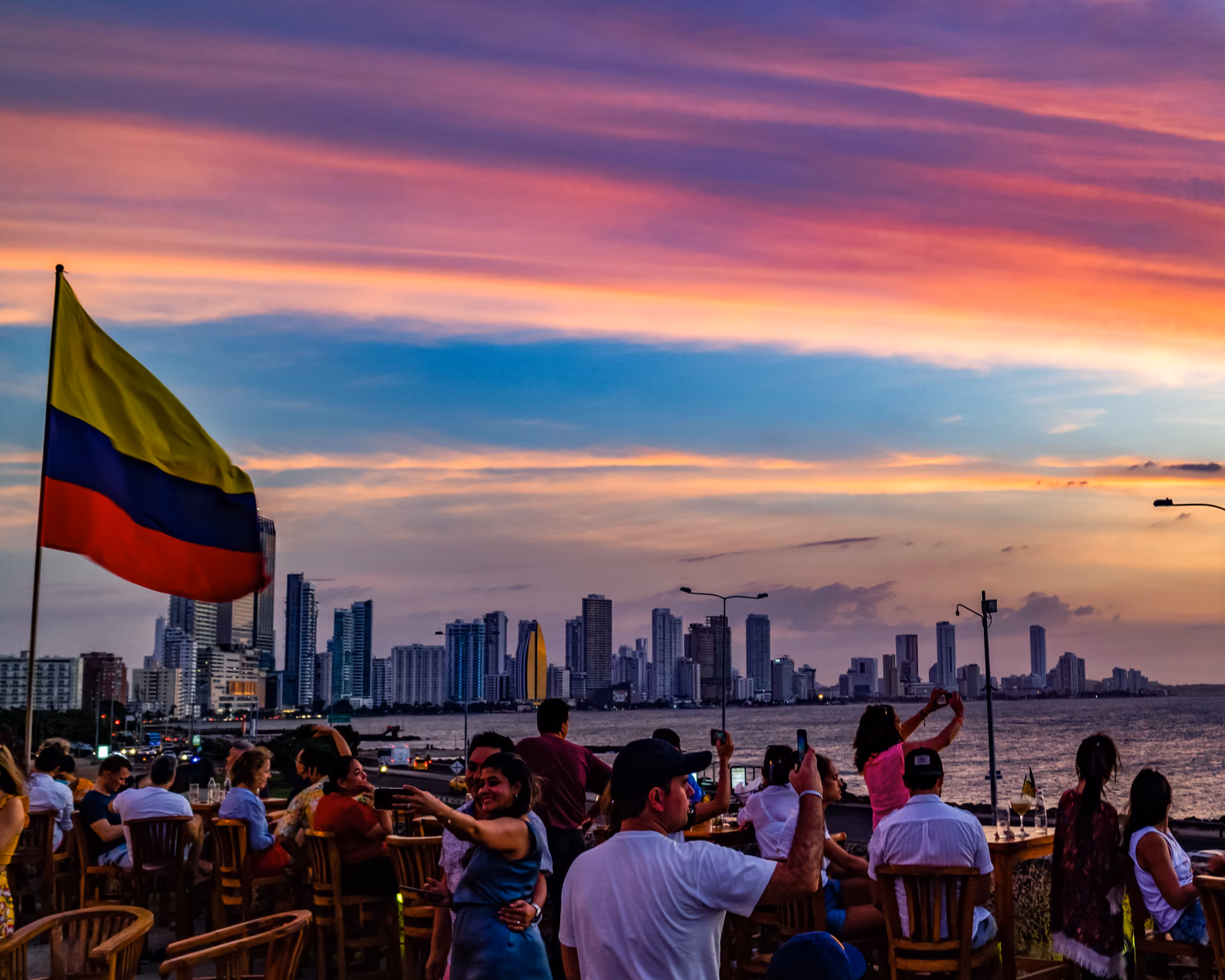 People at an outdoor venue with a Colombian flag and city skyline by the water during a colorful sunset.