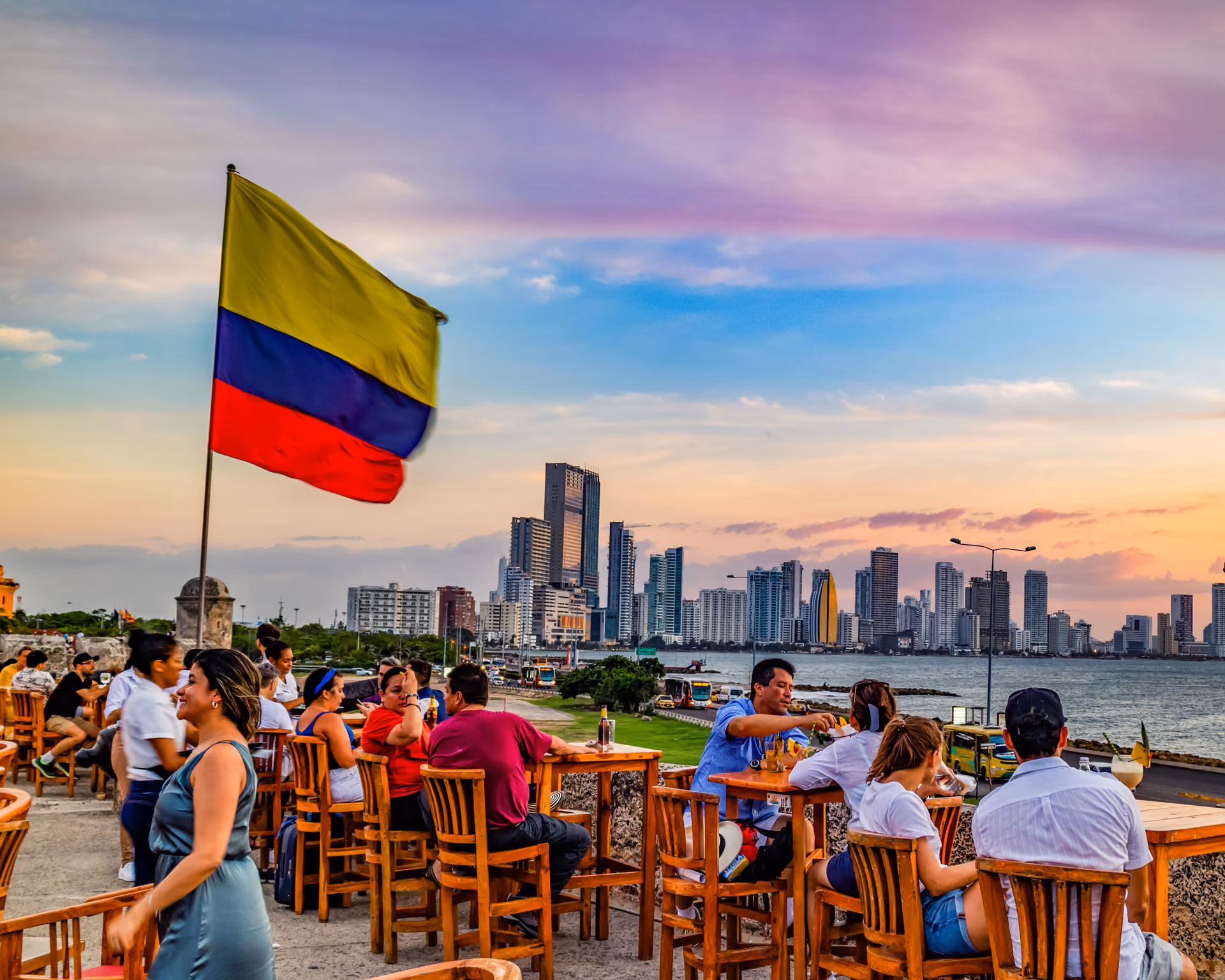People dining outdoors near water with Colombian flag and city skyline at sunset in the background.