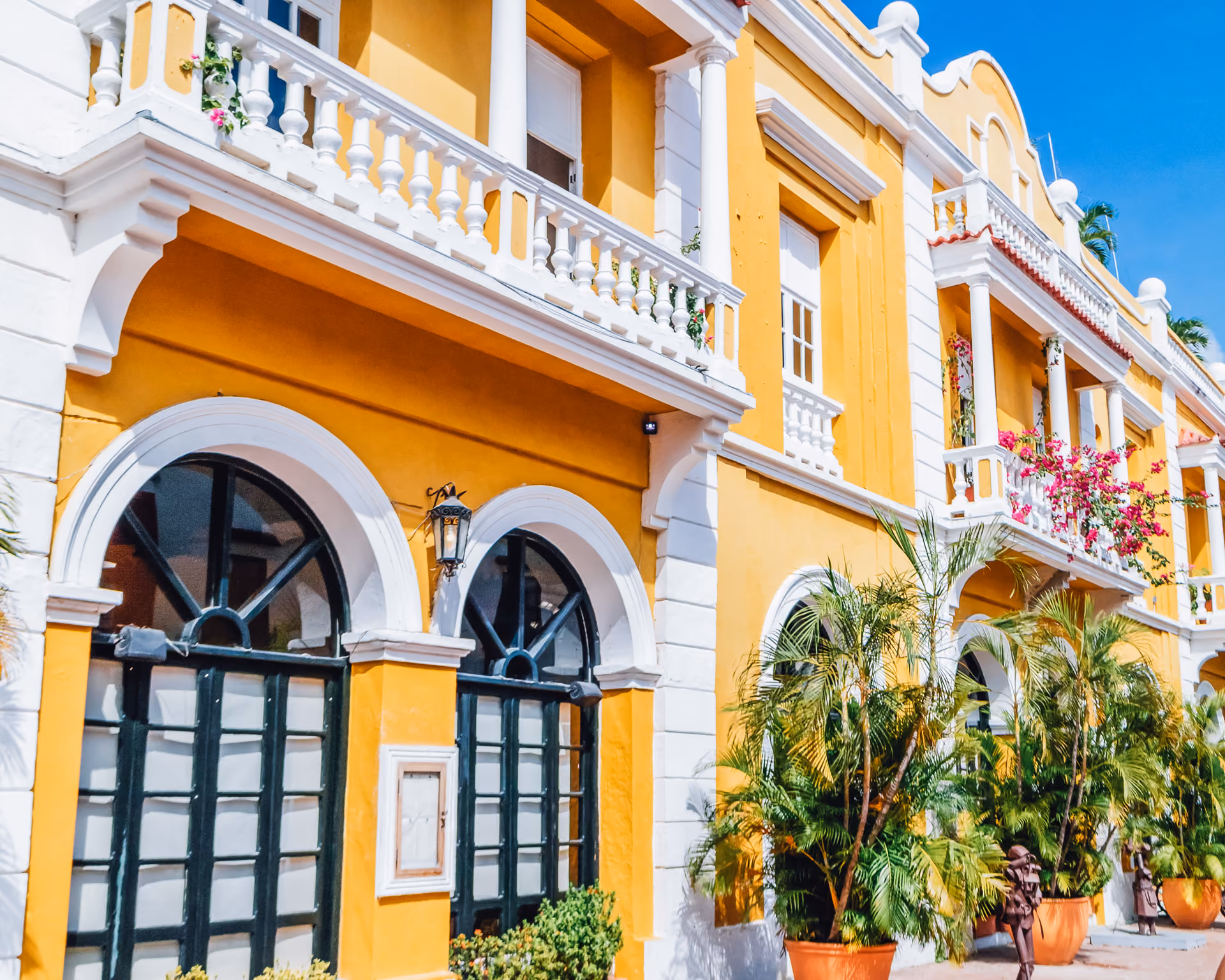 Yellow building with white trim, arched windows and doors, potted palm plants, and balconies with flowers.