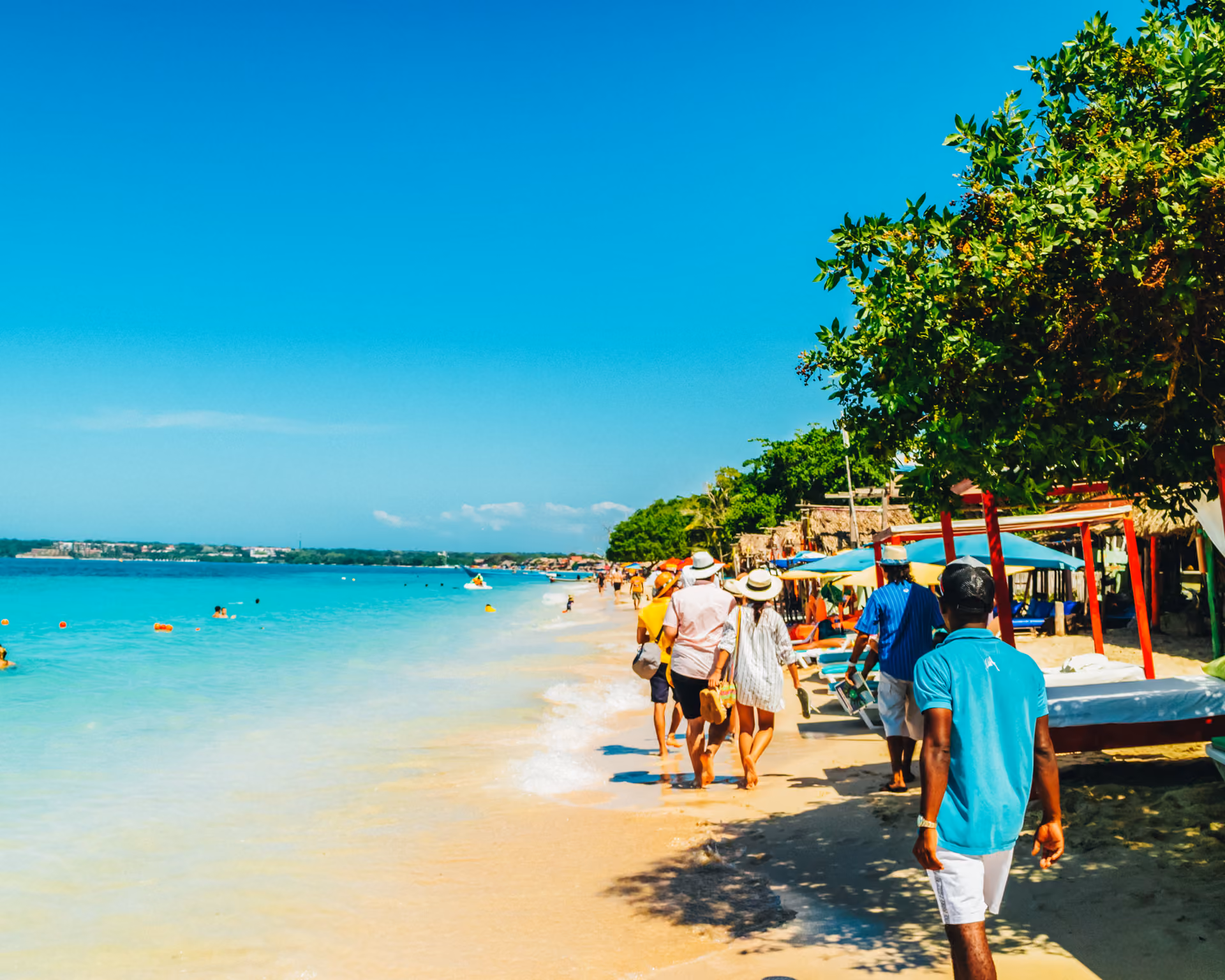 People walking along a sunny beach with clear blue water and shaded lounge areas under trees.