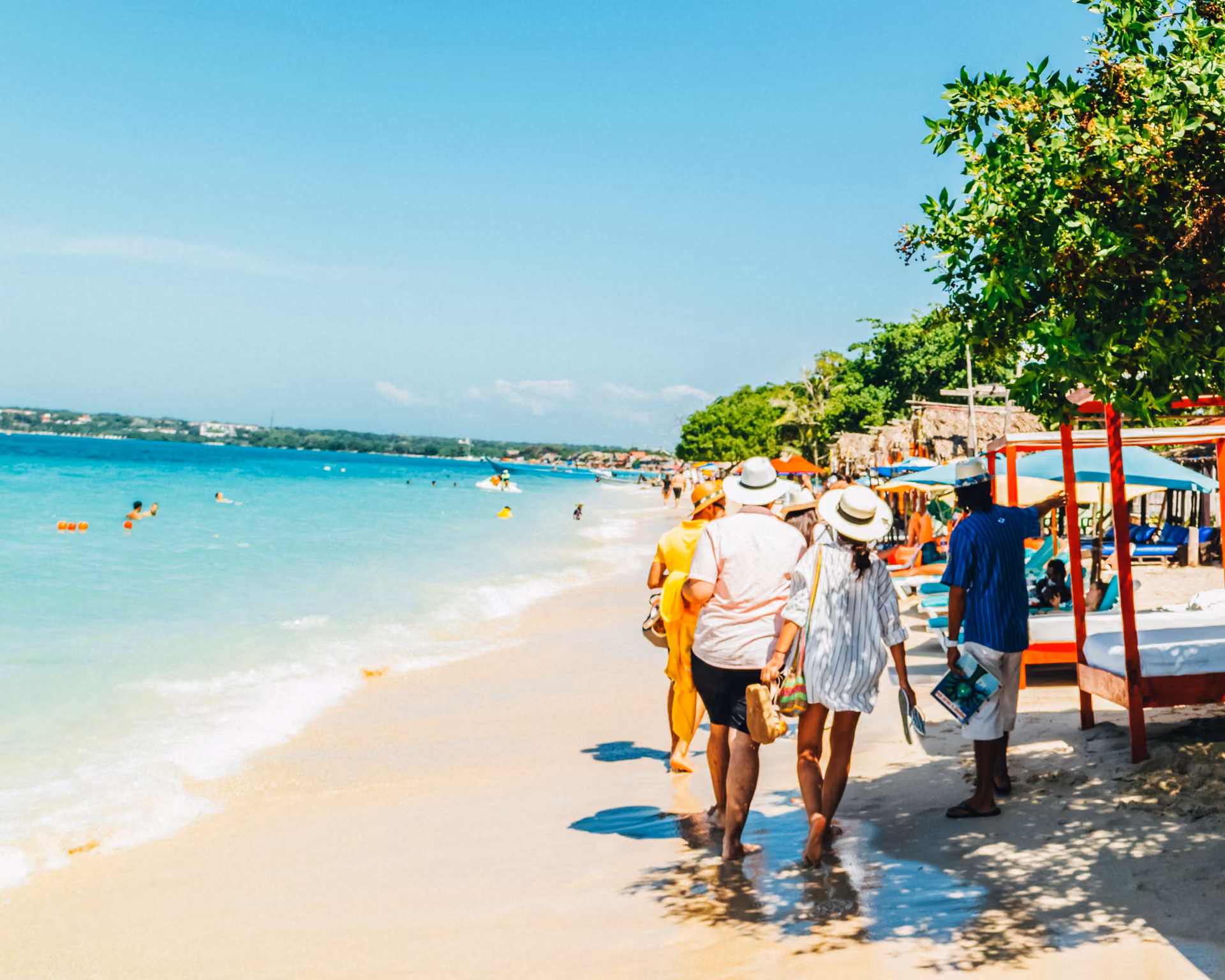 People walking along a sunny beach with turquoise water and shaded lounge beds.