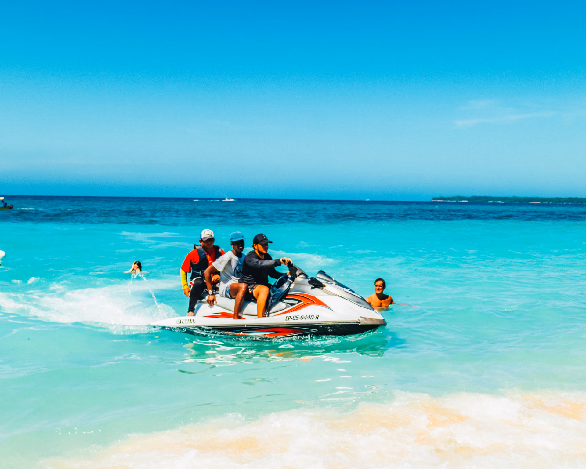 Three men riding a jet ski in bright turquoise ocean near the sandy shore under a clear sky.