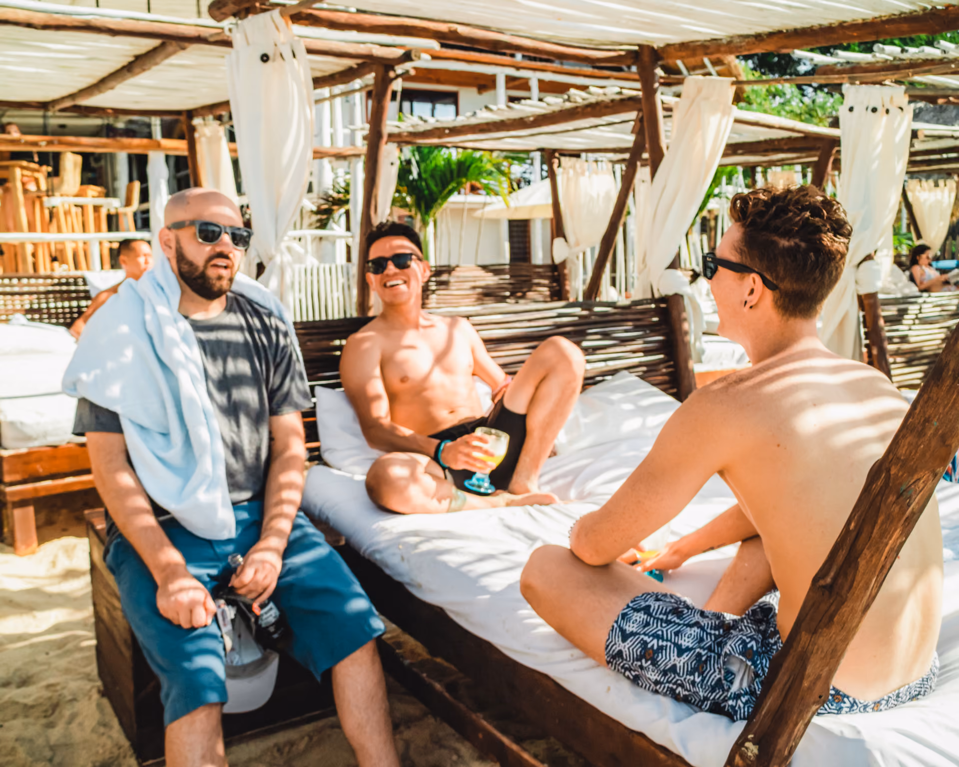 Three men wearing sunglasses relaxing on a shaded beach bed, one holding a drink, in a sunny setting.