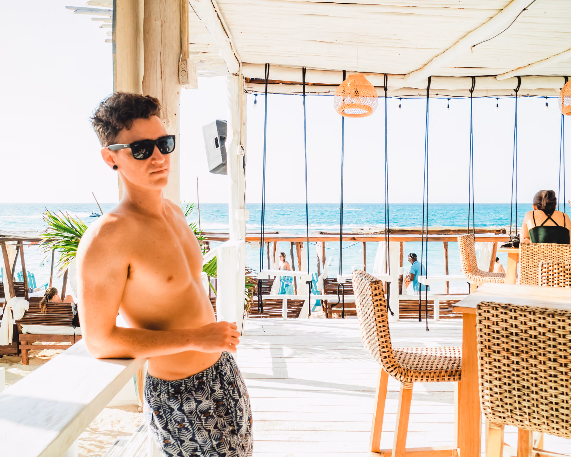 Shirtless man with sunglasses leaning on white railing at a beachside bar with ocean view.