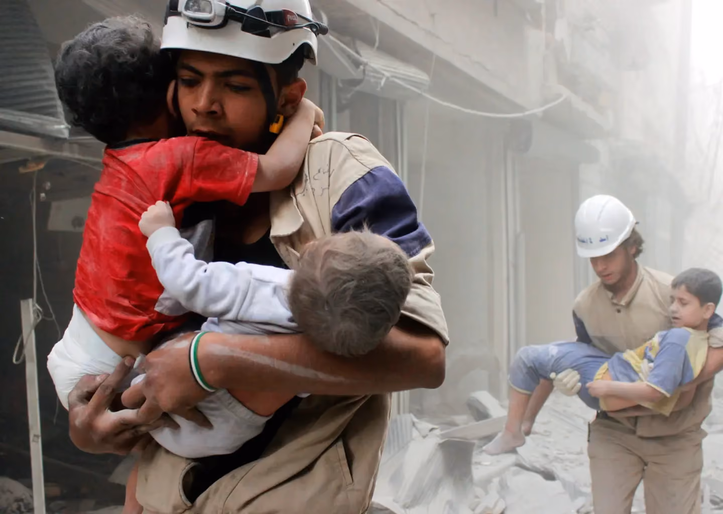 Rescue workers evacuating children from a disaster-stricken area amid rubble and dust.