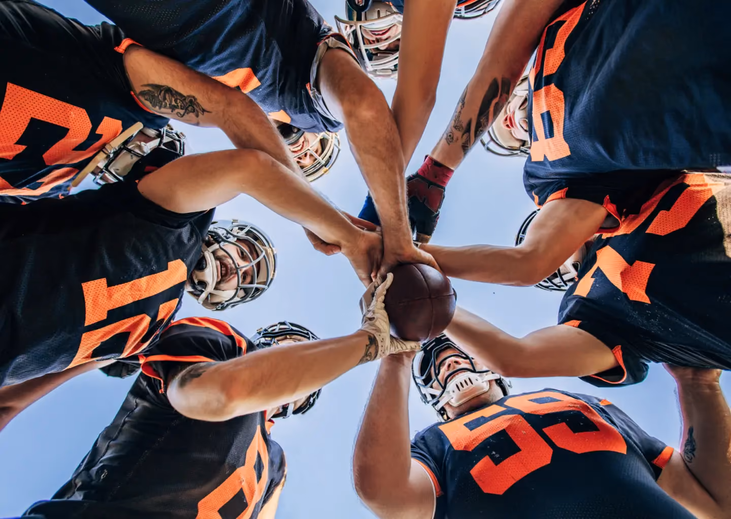 Low-angle view of American football players in uniform gathering in a team huddle, joining hands around a football before competition.
