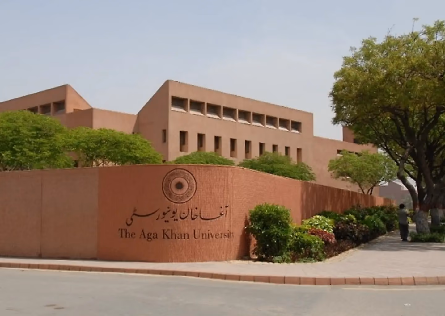 Main entrance to The Aga Khan University campus, showing the university sign and tree-lined walkways.
