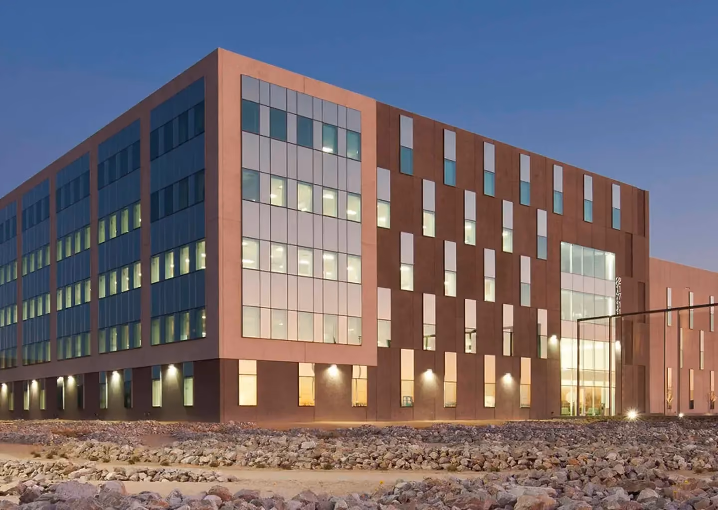 Exterior view of a modern corporate office building featuring large glass windows, architectural detailing, and illuminated workspaces at dusk.