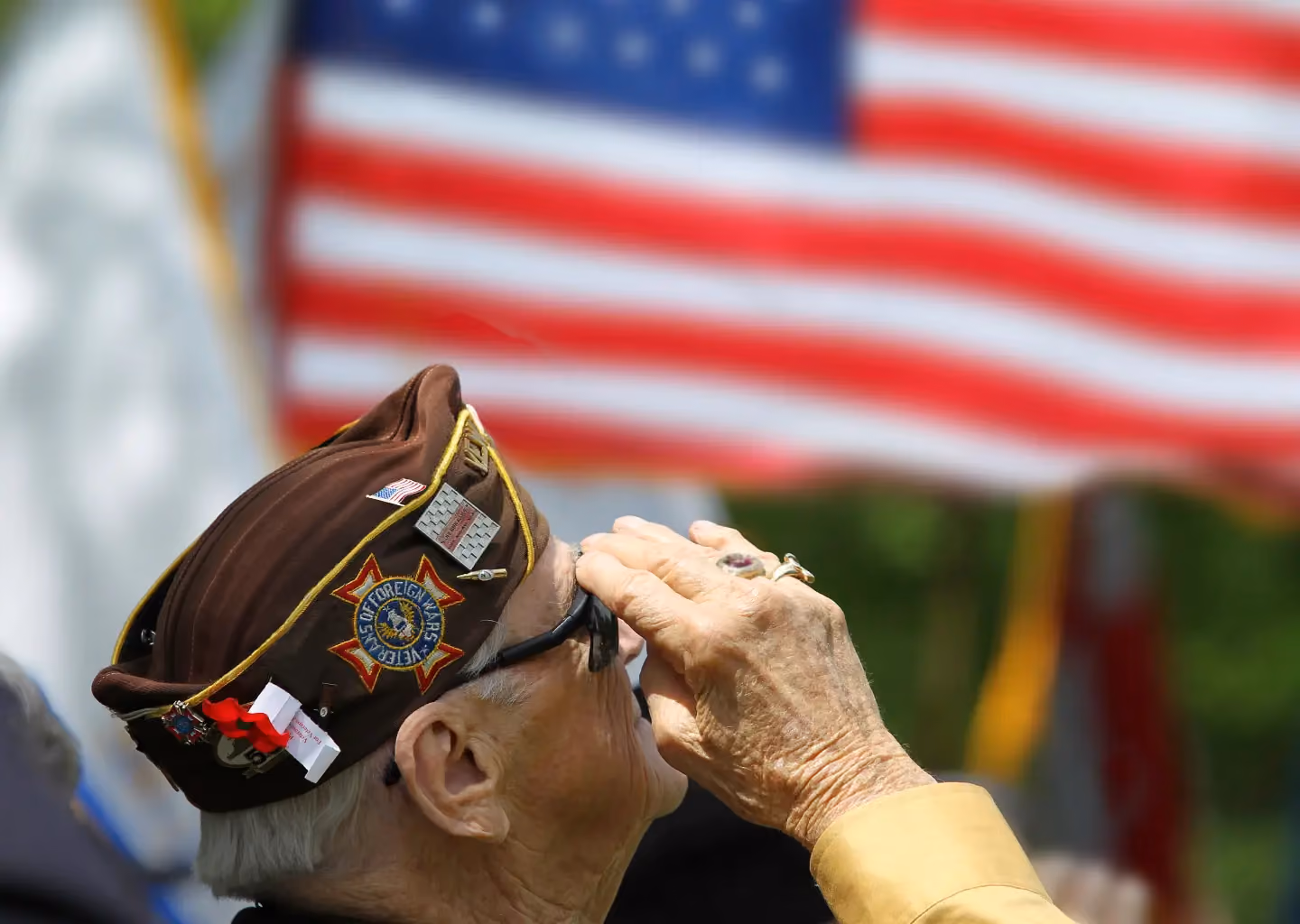 Elderly military veteran wearing a VFW cap raises a salute while facing an American flag.