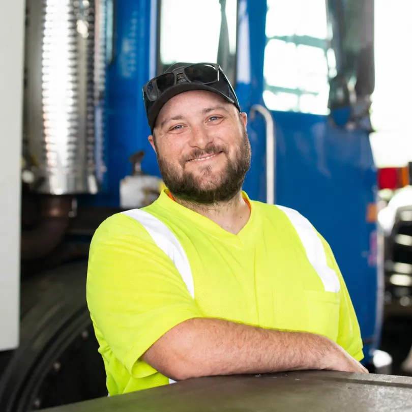 Smiling man in a yellow safety shirt and black cap standing in front of a blue truck.