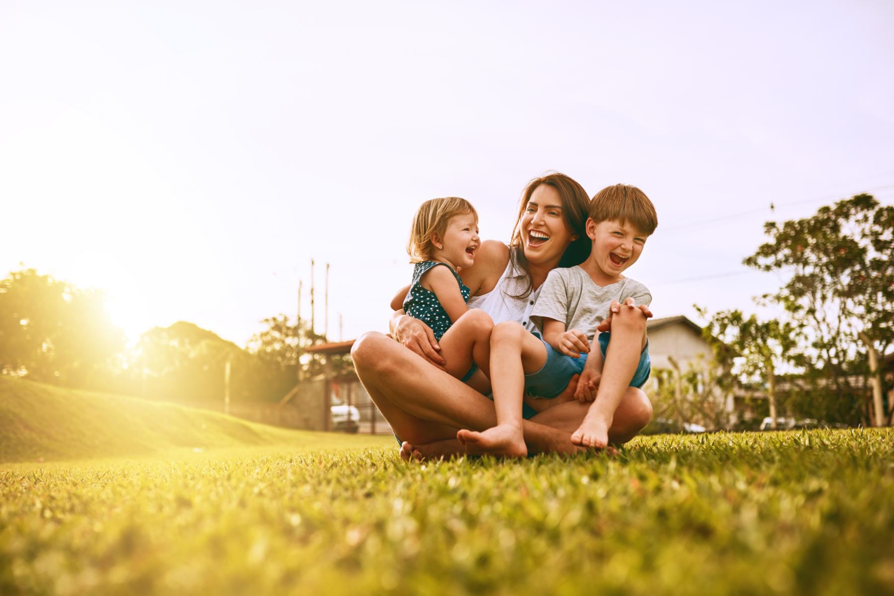 Smiling mother sitting on grass outdoors holding laughing young boy and toddler girl in sunset light.