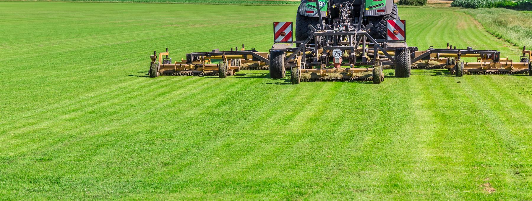 Large tractor with wide mower attachment cutting fresh green grass on a vast field.