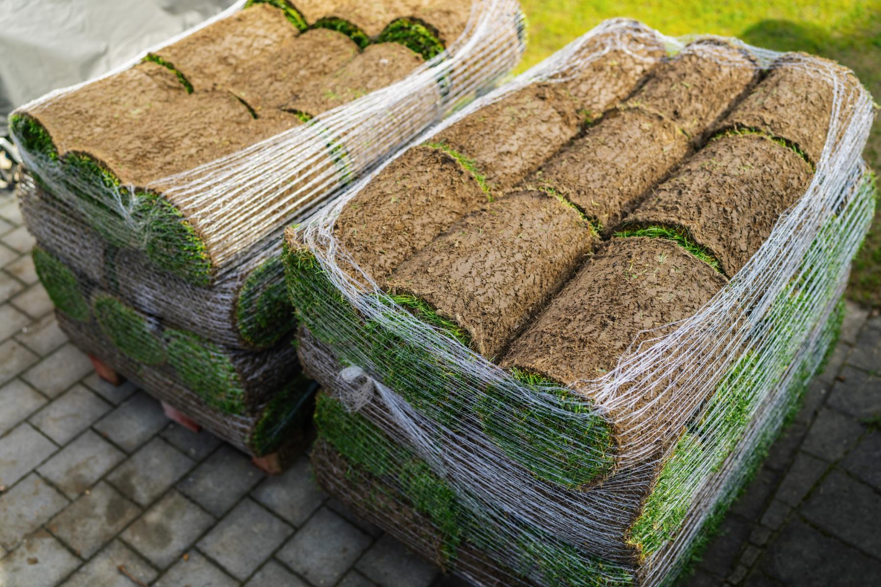Two pallets stacked with rolled sod wrapped in white plastic netting on a paved surface.