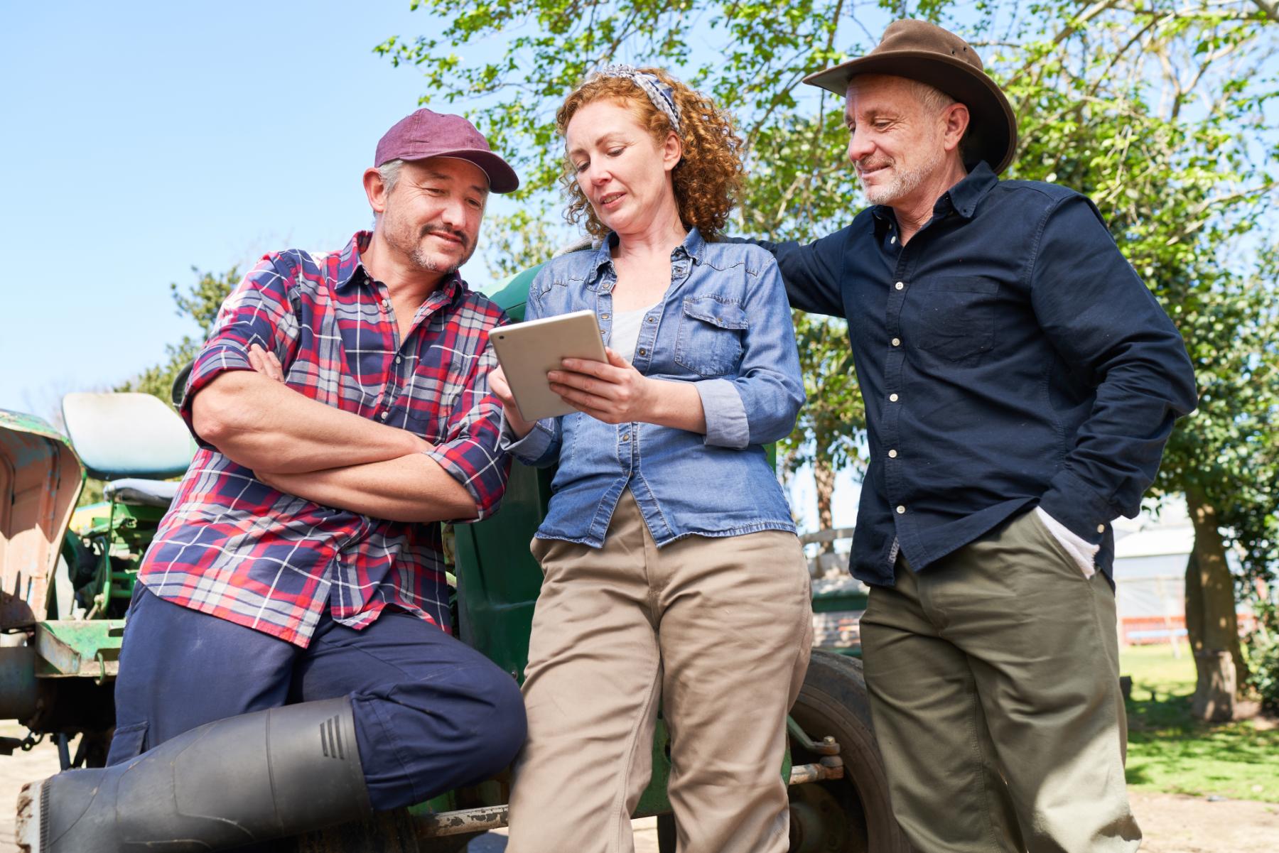 Two men and a woman standing by a tractor outdoors, the woman showing tablet content to them.