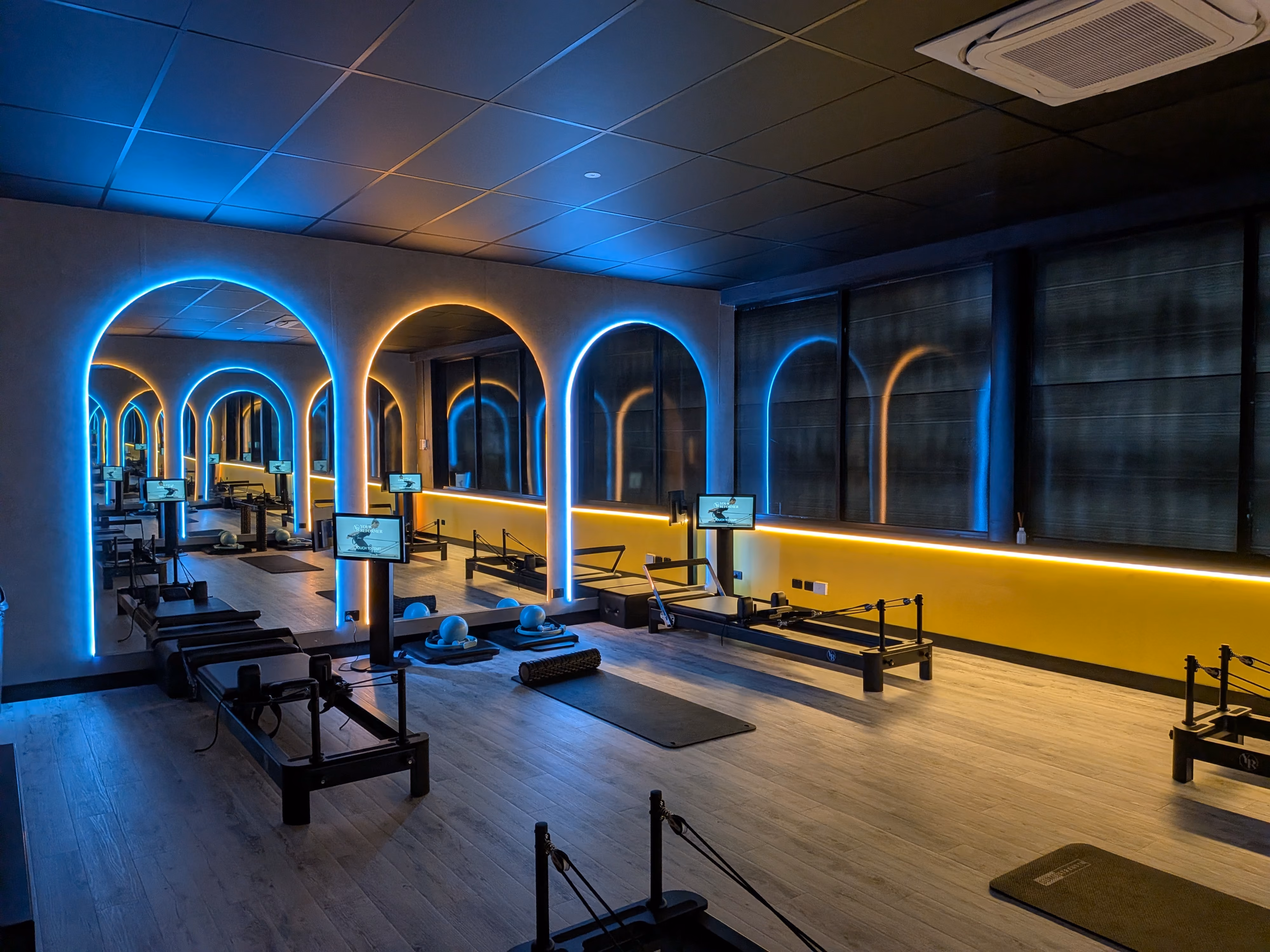 Modern Pilates studio with reformer machines, blue and yellow LED arch lighting, and wood flooring.