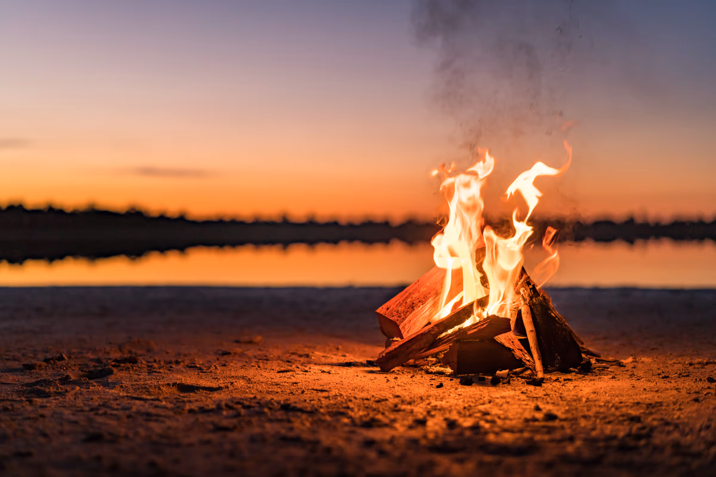 Vuur brandt op het zand bij zonsondergang aan het water met een vage horizon op de achtergrond.
