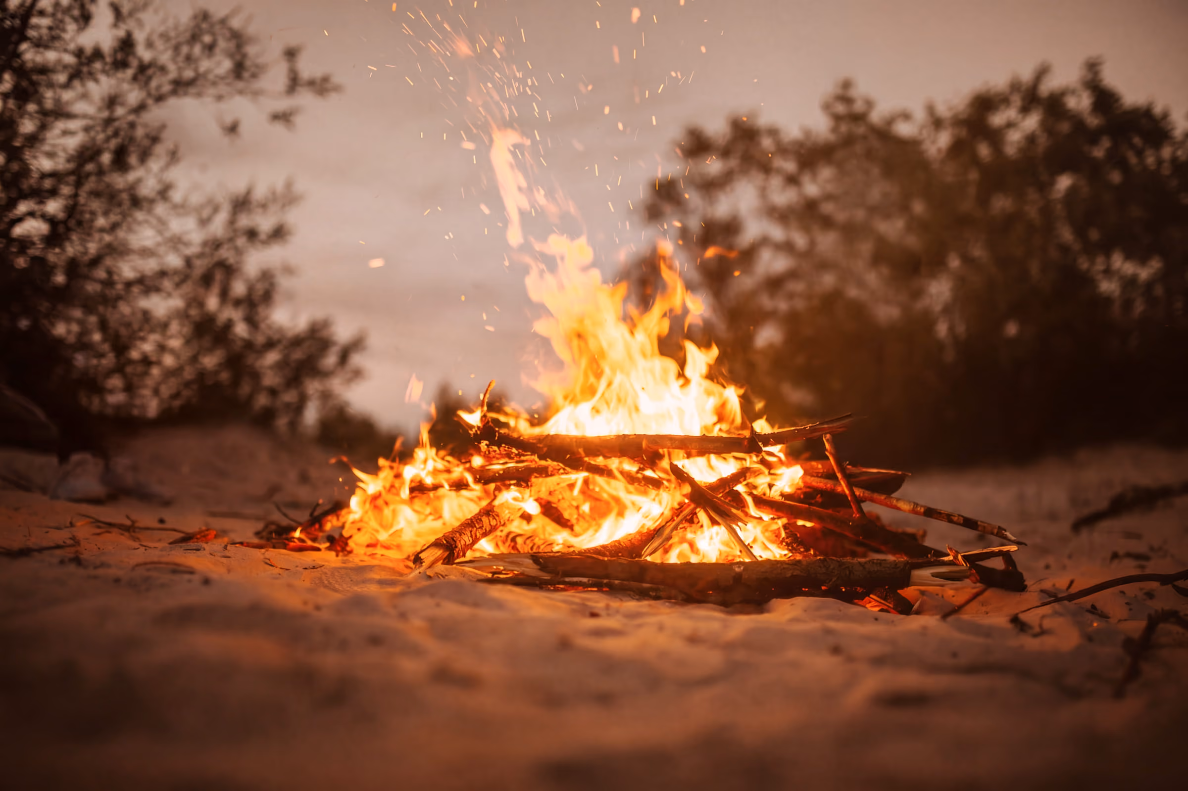 Een kampvuur brandt op zand met vonken en bomen op de achtergrond bij schemering.