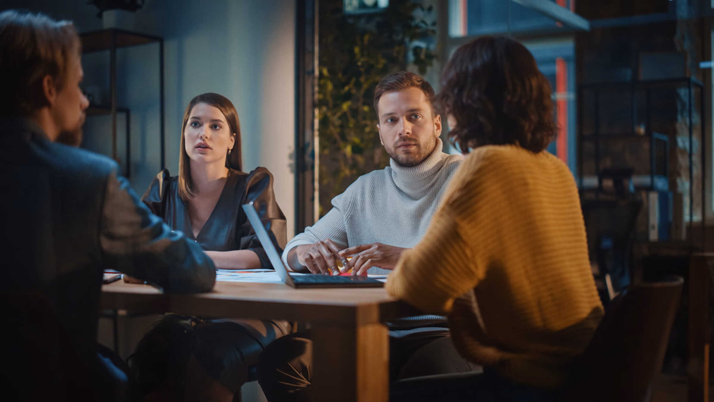Vier collega's zitten 's avonds aan tafel in gesprek, met een gespannen sfeer.