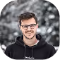 Young man with glasses smiling, wearing black hoodie, blurred outdoor background.