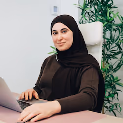 Une femme portant un hijab noir, assise à un bureau avec un ordinateur portable, sourit doucement.