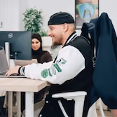 Un homme souriant en veste et bonnet travaille sur un ordinateur avec une veste Dishop, une femme voilée le regarde en arrière.