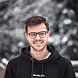 Jeune homme souriant portant des lunettes et un sweat à capuche noir sur fond flou extérieur.