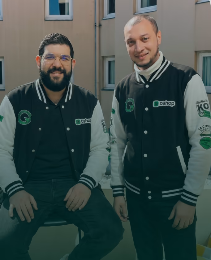 Deux hommes portant des vestes noires et blanches avec logos posant devant un bâtiment beige.