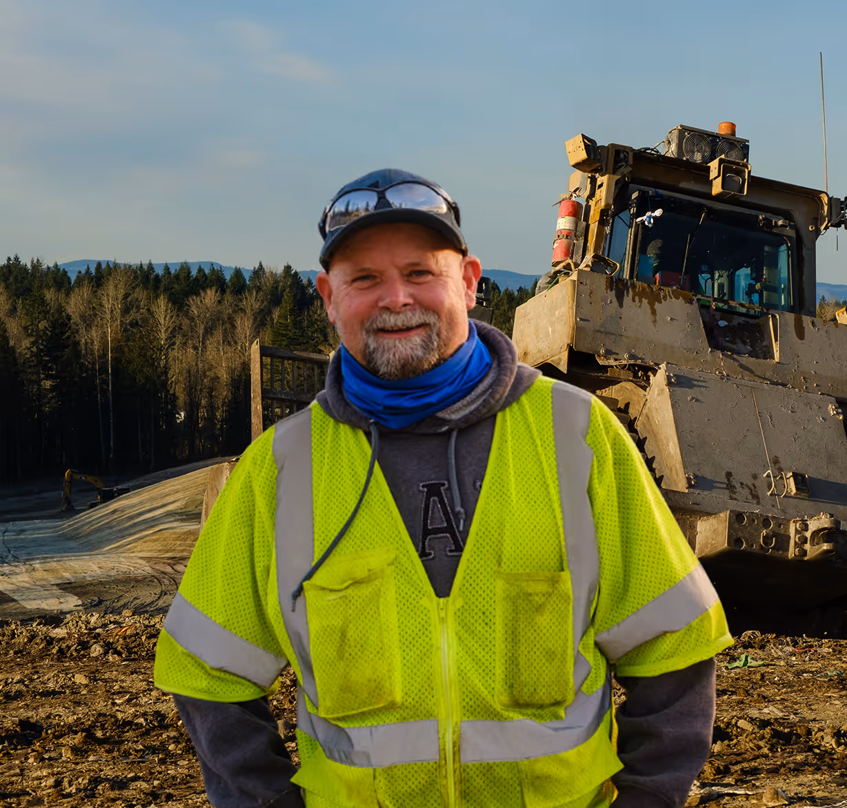 Construction worker in a yellow safety vest stands in front of a bulldozer at a dirt worksite.