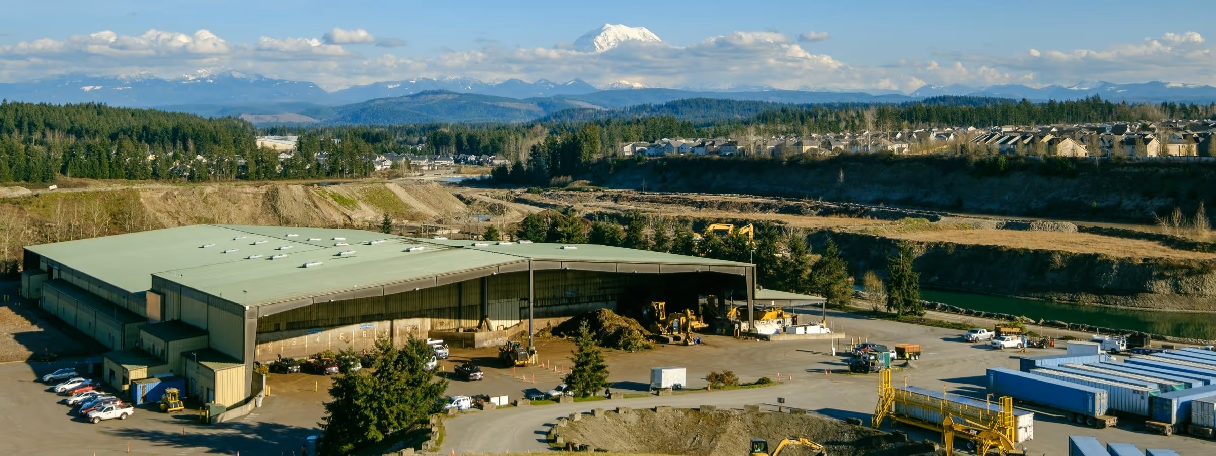 Aerial view of a recycling facility with trucks, vehicles, trees, and snowy mountains in the background.