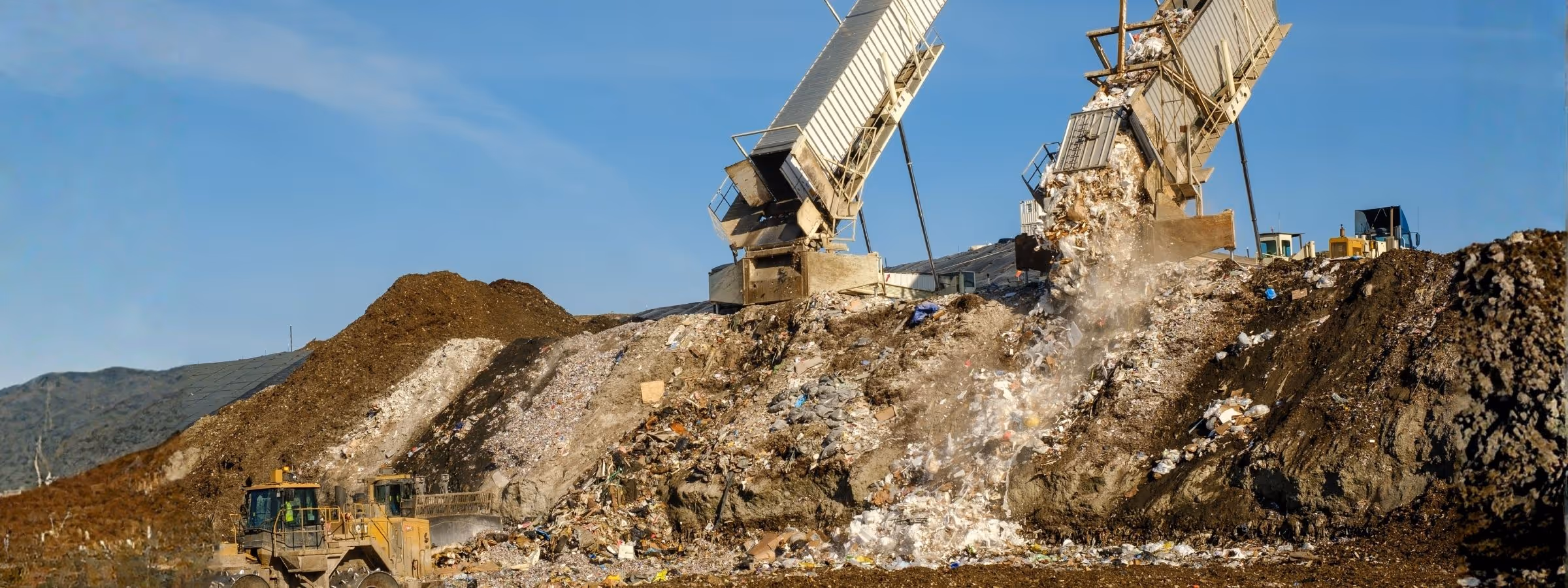 Dump trucks unloading waste onto large landfill piles with clear sky in the background.