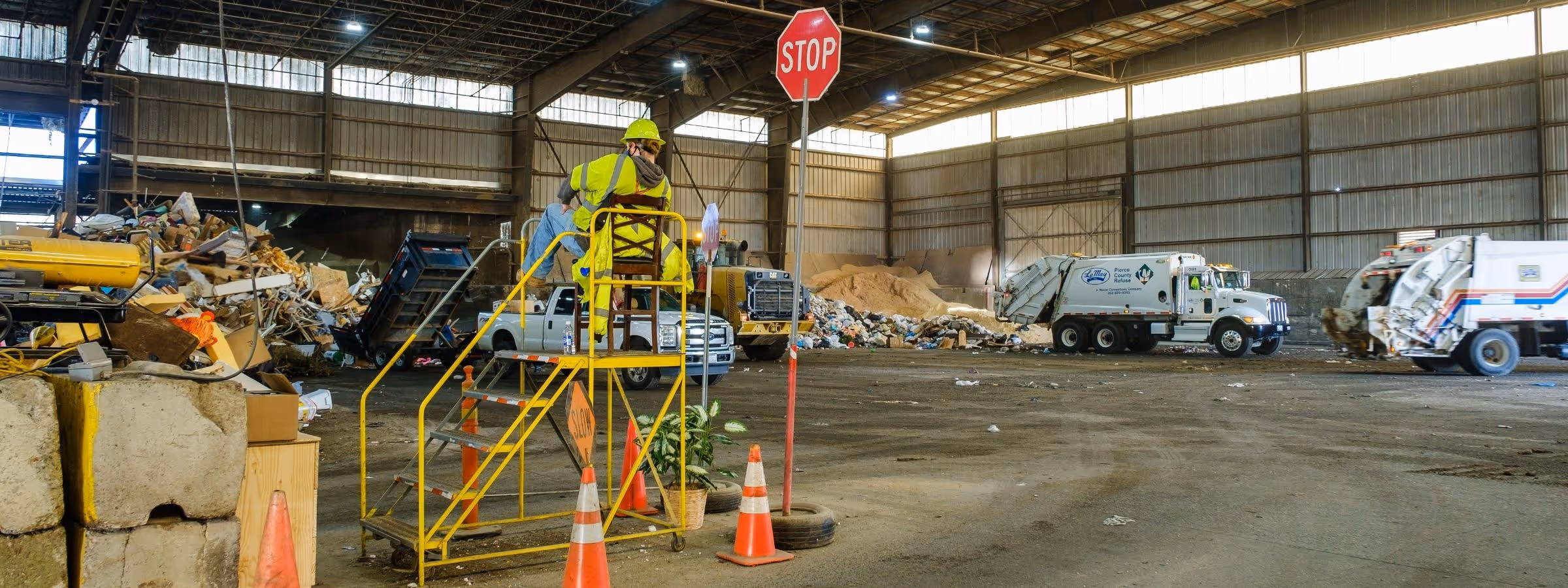 Worker in yellow safety gear on platform with stop sign at recycling transfer station with trucks and waste piles.